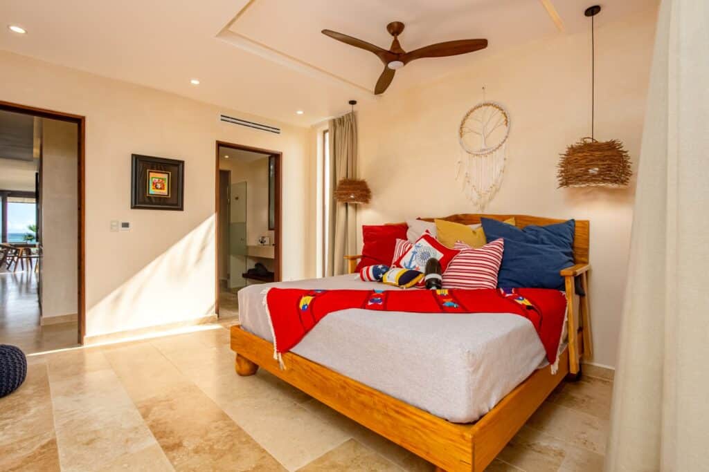 Bedroom with wooden bed frame, colorful pillows, a dreamcatcher on the wall, and natural light in a beachfront condo in Tulum at Kaoba Beachfront Condo.