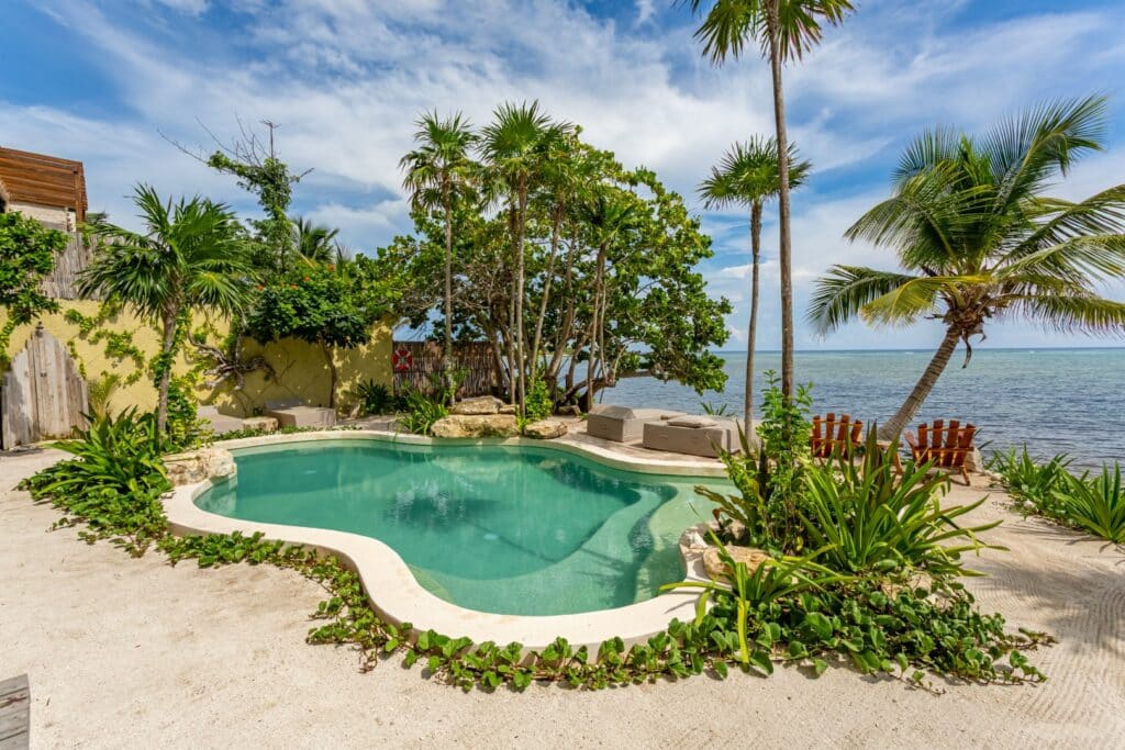 View of a tropical backyard with a private pool, sand, lush green trees, and the ocean in the background at beachfront in Tulum at Kaoba Beachfront Condo.