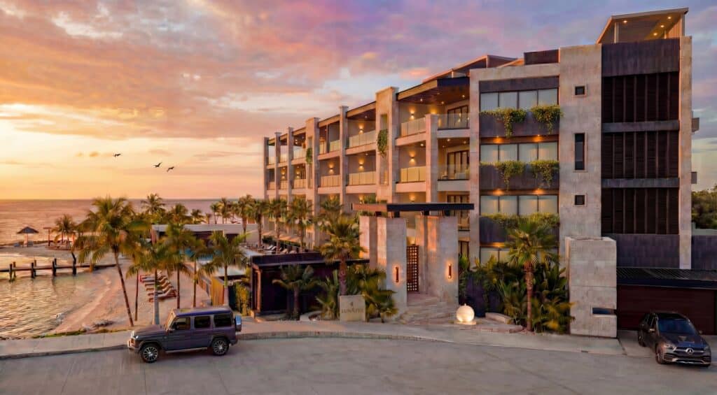 Modern multi-story building with balconies near a beach, surrounded by palm trees during sunset at Luna Azul 101 in Bahia Xcacel, Puerto Aventuras.