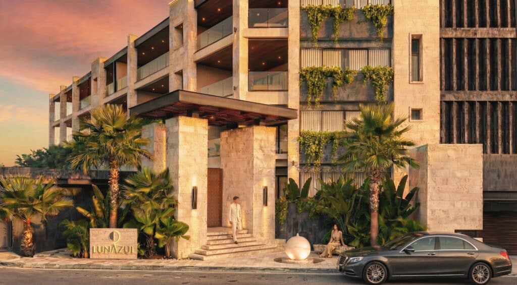 Front view of a modern multi-story building with glass balconies and lush greenery, with a sunset sky at Luna Azul 103 in Bahia Xcacel, Puerto Aventuras.