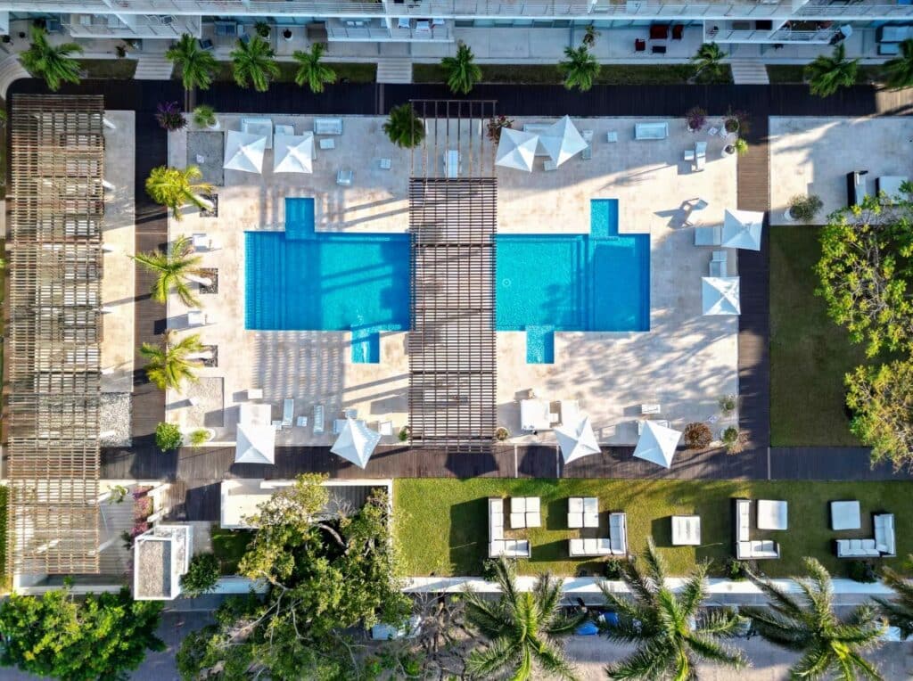 Aerial view of a swimming pool area at Magia 202 F in Playa Centro, Playa del Carmen, with sun umbrellas, lounge chairs, palm trees, and surrounding landscaping.