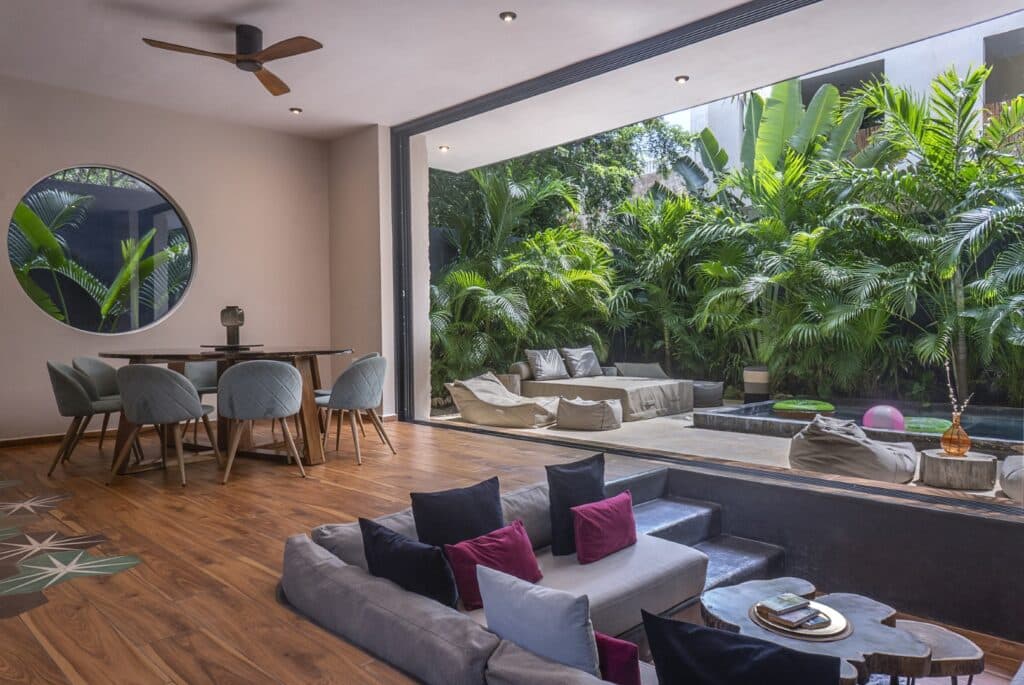 Modern living room with floor-to-ceiling sliding glass doors opening to a lush tropical garden and pool area in Tulum.