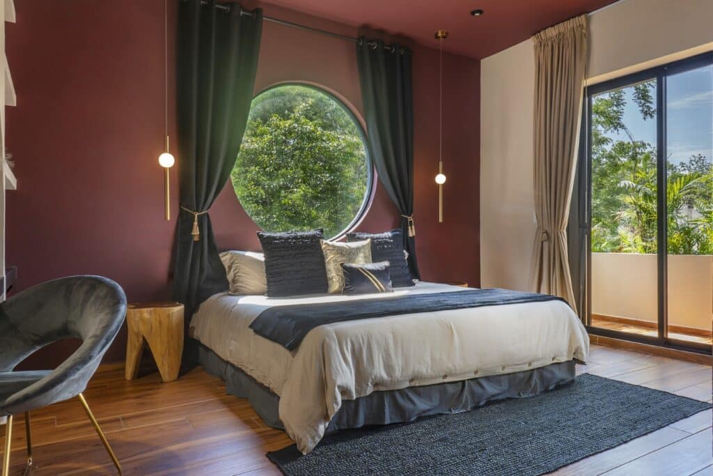 Interior bedroom with a round window, dark green curtains, and a sliding glass door opening to greenery in Tulum's Region 15.