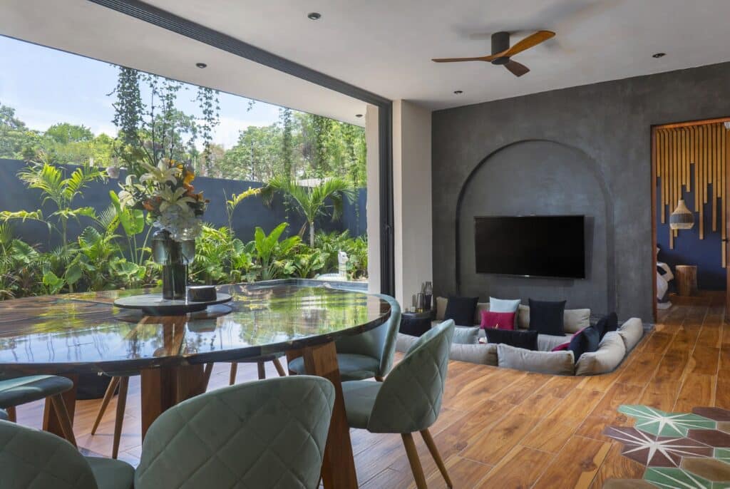 Interior view of a modern Tulum residence with a living area featuring a dark accent wall, large window overlooking lush greenery, and wooden flooring.