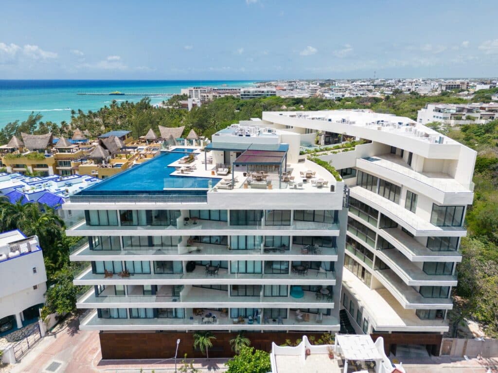 A modern multi-story condominium building with large glass windows, balconies, and a rooftop pool overlooking the ocean and cityscape in Playa del Carmen at Marila Condo.