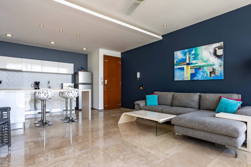 Interior of a modern living room with a gray sofa, colorful pillows, blue accent wall, and abstract artwork at Coco Beach, Playa del Carmen at Menesse 301 2-Bedroom Condo.