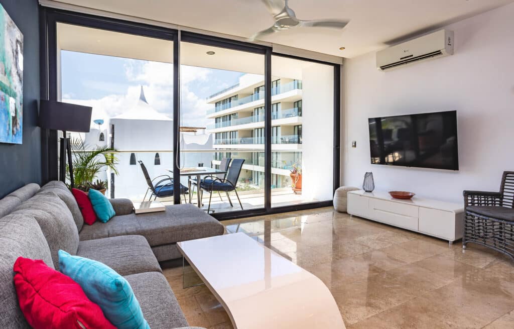 Living room with large glass doors opening to a balcony overlooking neighboring modern white buildings and a blue sky at Menesse 301 2-Bedroom Condo in Coco Beach, Playa del Carmen.