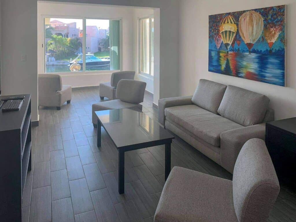 Living room with gray sofa, armchairs, a large window showing boats and water, and colorful abstract art at Nautica 8 Unit Marina Front Building in Boulevard Puerto Aventuras.