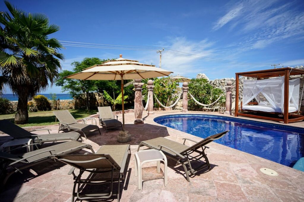 View of a swimming pool with lounge chairs, umbrellas, and a poolside cabana in a tropical outdoor setting at Isla Punta Sur, Isla Mujeres at Casa de las Cúpulas Ocean View Villa.