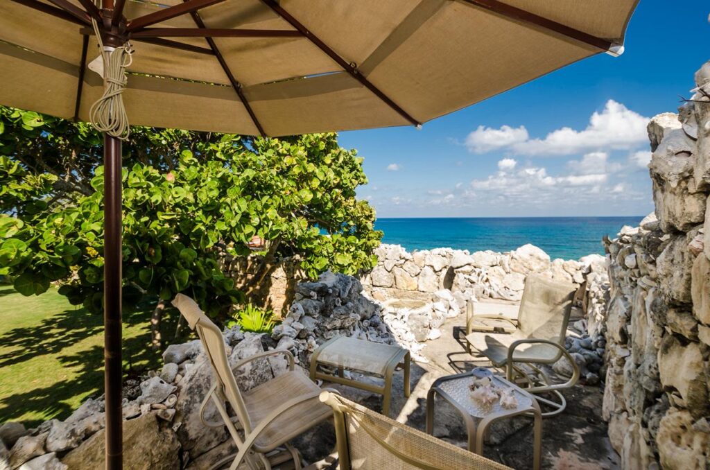 View of the ocean through a stone wall with outdoor lounge chairs and an umbrella at Casa de las Cúpulas Ocean View Villa in Isla Mujeres.