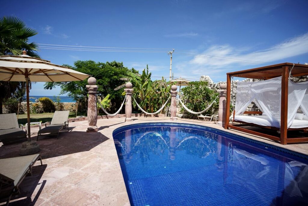 Ocean view swimming pool area with a shaded cabana, umbrellas, lounge chairs, and lush greenery at Casa de las Cúpulas in Isla Mujeres at Casa de las Cúpulas Ocean View Villa.