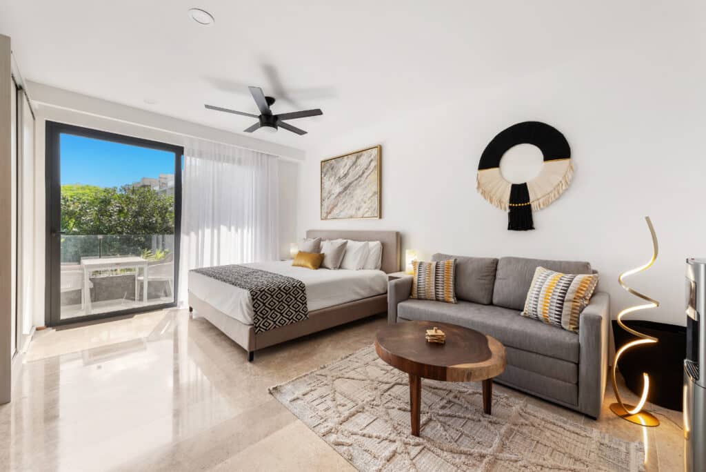 Living room with sliding glass door opening to balcony, featuring a bed, gray sofa with patterned pillows, and modern decor in bright space at Sea Tower Studio.