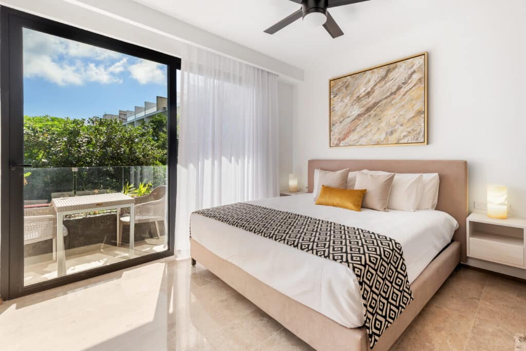 Bedroom with a bed, bedside tables, artwork, and sliding glass door leading to a balcony with greenery view in Playa del Carmen at Sea Tower Studio.