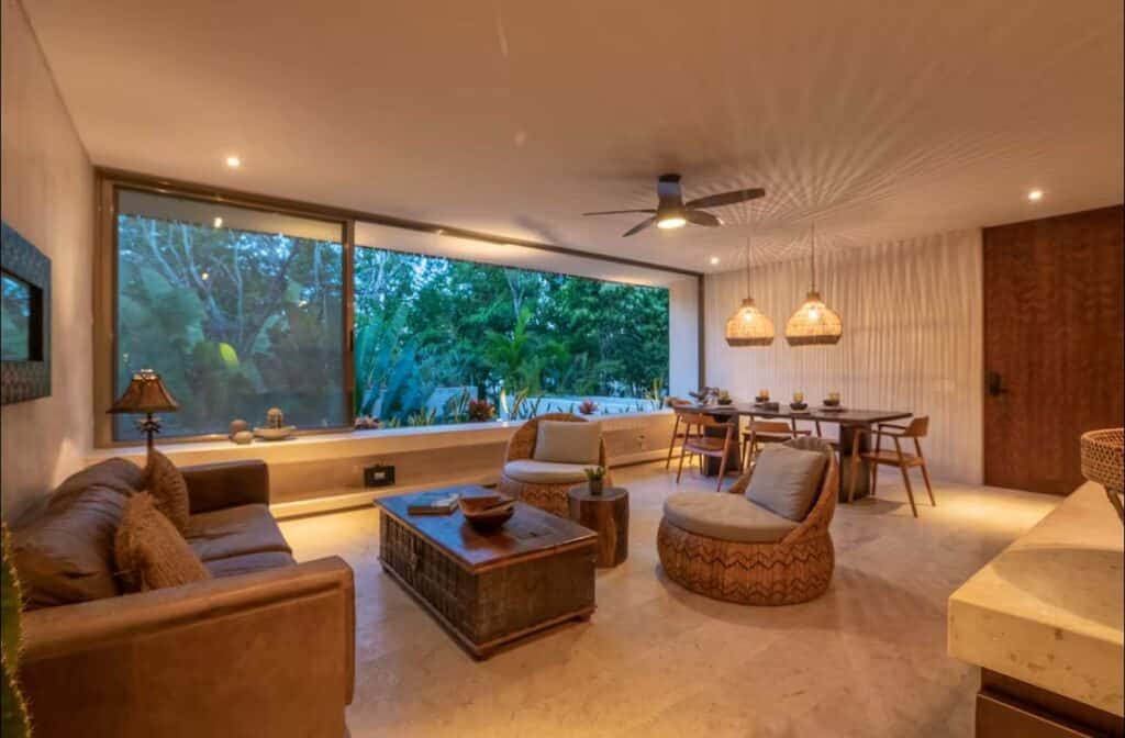 Interior view of Sky Jungle 207 in Tulum, featuring a spacious living room with a large window overlooking lush greenery, wood accents, and cozy lighting.
