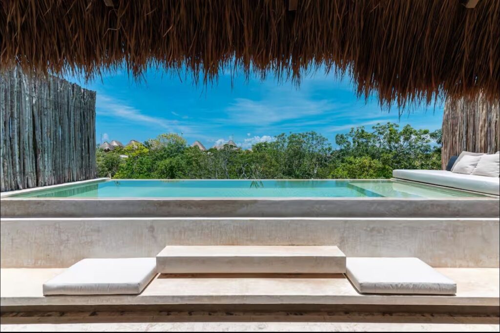 Sky Jungle 207 in Tulum features an infinity pool with a thatched roof, wooden privacy fencing, and lush green trees in the background under a blue sky.