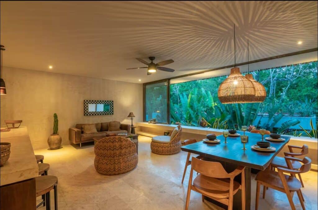 Interior living room area in Sky Jungle 207, Tulum, featuring a large window with lush greenery view, wooden dining table, rattan pendant light, and ceiling fan.