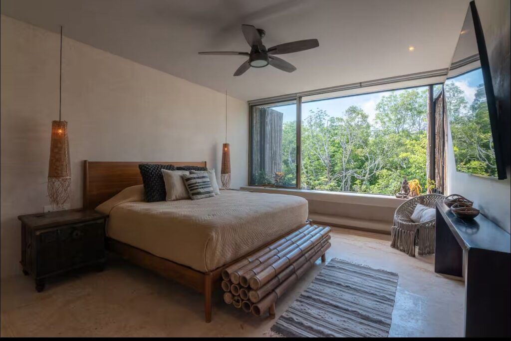 Interior view of a bedroom in Sky Jungle 207, Tulum, featuring a large window showing lush green trees and a ceiling fan.