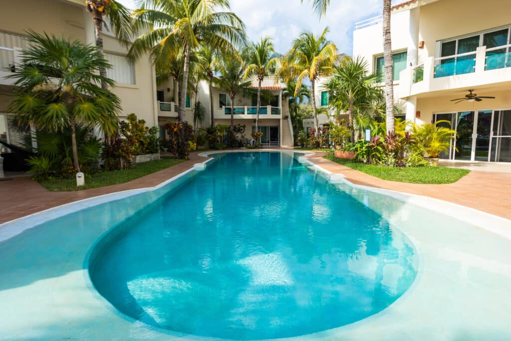 View of a curved swimming pool surrounded by tropical palm trees and greenery at a modern residential complex in Puerto Aventuras at Sotavento Penthouse.