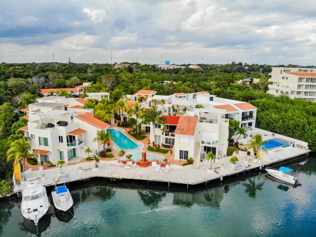 Luxury white residential complex with red-tile roofs, surrounded by lush green trees, overlooking a marina with boats and water in Marina Front, Puerto Aventuras.