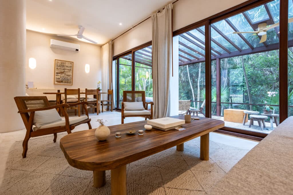 Living room with large glass doors opening to lush greenery, wooden furniture, and neutral decor in Tao Ocean D1 at Tao Ocean D1 3-Bedroom House in Akumal.