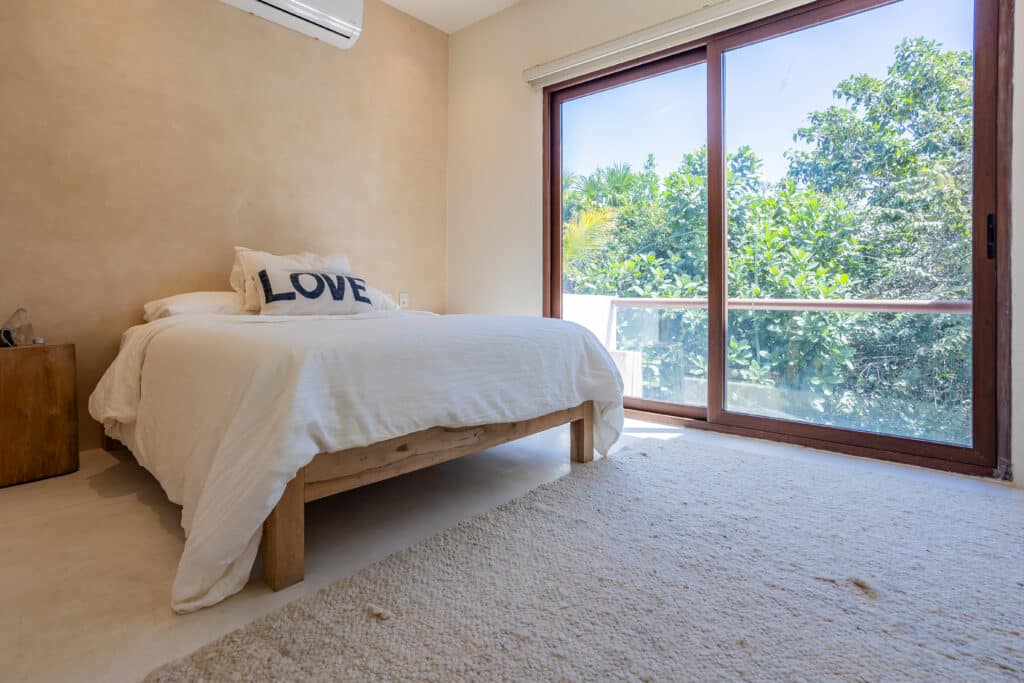 Bedroom with a wooden bed frame, white bedding, a decorative pillow reading 'LOVE', and large glass sliding doors showing lush green trees at Tao Ocean D1 3-Bedroom House in Akumal.