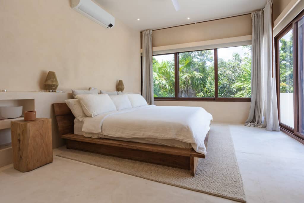 Bedroom with large windows showing lush greenery, white bedding, wooden bed frame, and modern decor at Tao Ocean D1 3-Bedroom House in Akumal.