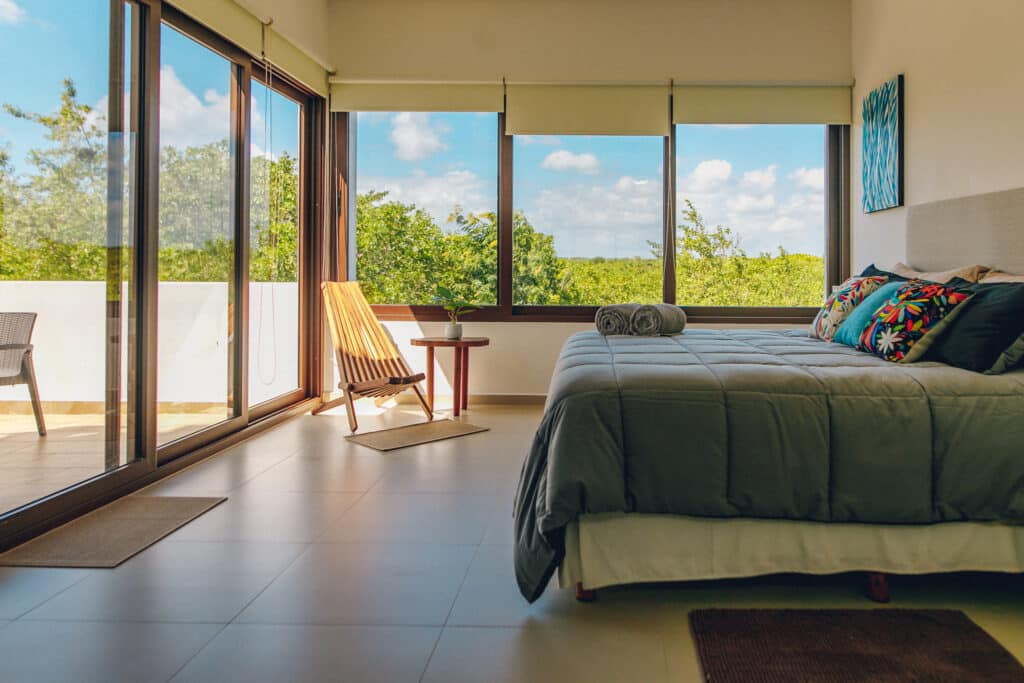 Bedroom with large windows overlooking lush green trees and a blue sky in the background, featuring a bed with colorful pillows, a small side table, and a wooden chair in Akumal.