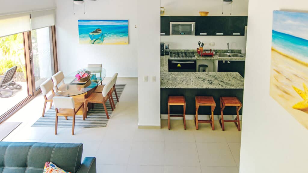 Interior view of a dining area and kitchen with modern furnishings, a sliding glass door opening to an outdoor patio, and colorful artwork at Tao Ocean H2 3-Bedroom House in Akumal.