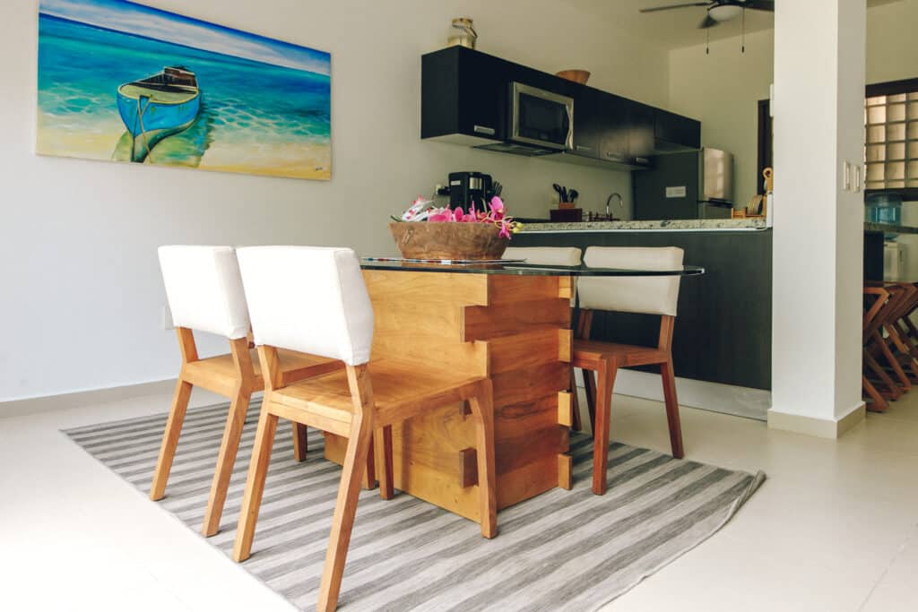 Modern dining area with wooden table and white chairs in an open-concept kitchen with black cabinetry and a colorful boat painting at Tao Ocean H2 3-Bedroom House in Akumal.