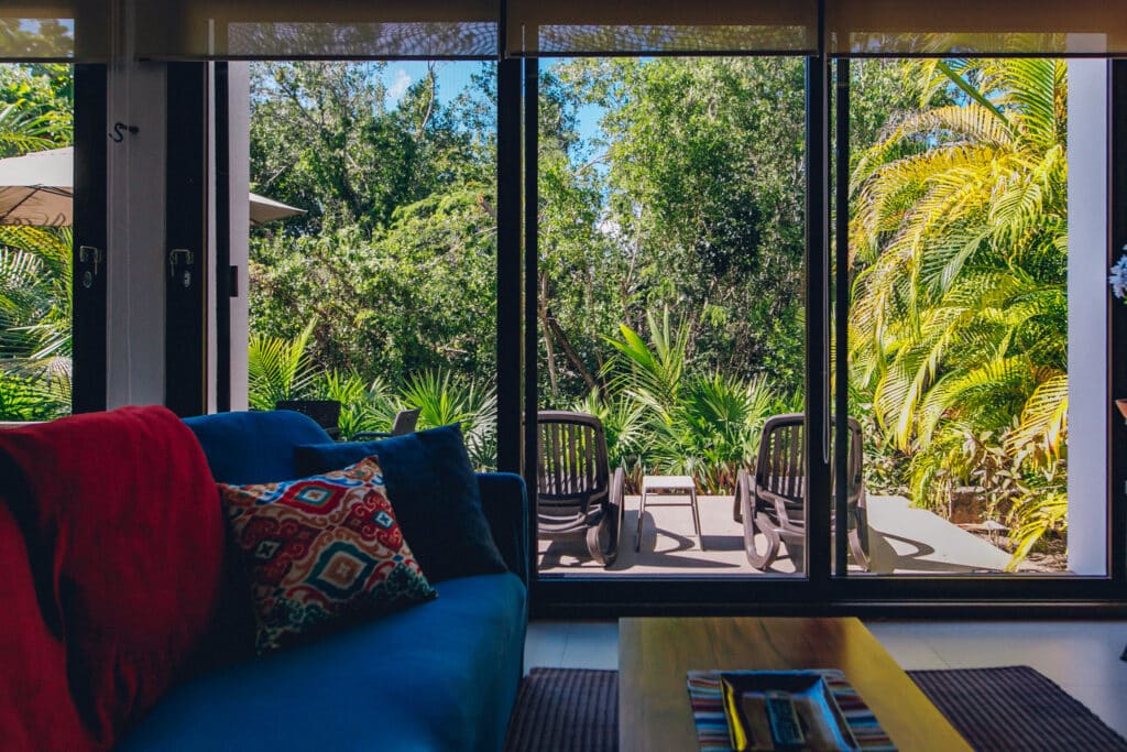 Living room with large windows overlooking lush greenery in jungle, Akumal, featuring colorful pillows and a wooden coffee table at Tao Ocean H3 3-Bedroom House.