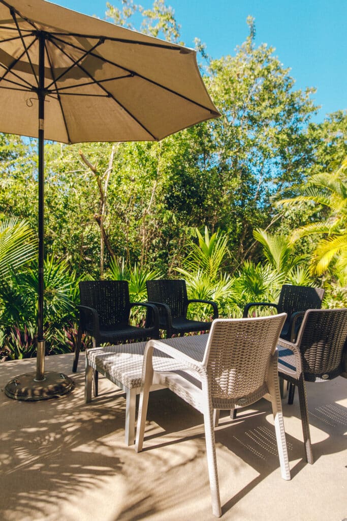 Outdoor seating area with white and black chairs, a large umbrella, surrounded by lush green plants and trees in the jungle near Akumal at Tao Ocean H3 3-Bedroom House.