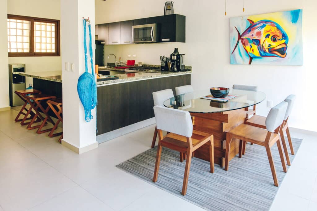Dining room with a glass table, six chairs, and a colorful abstract fish painting on the wall in a house surrounded by jungle in Akumal at Tao Ocean H3 3-Bedroom House.