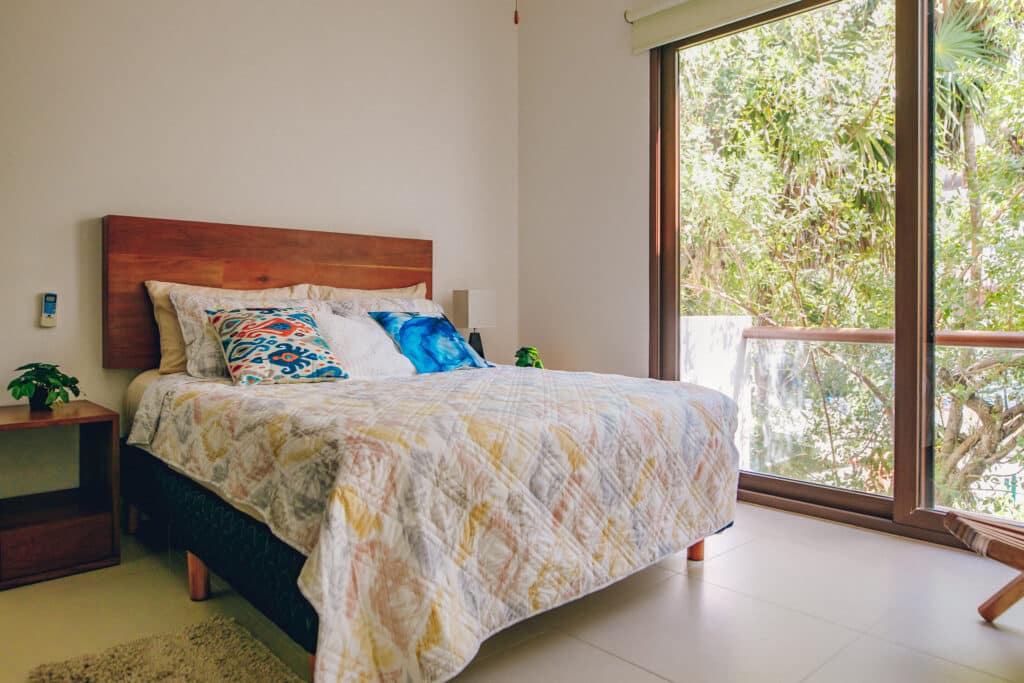 Bedroom with a wooden bed frame, colorful pillows, a nightstand, and a large sliding glass door opening to a lush jungle view in Akumal at Tao Ocean H3 3-Bedroom House.