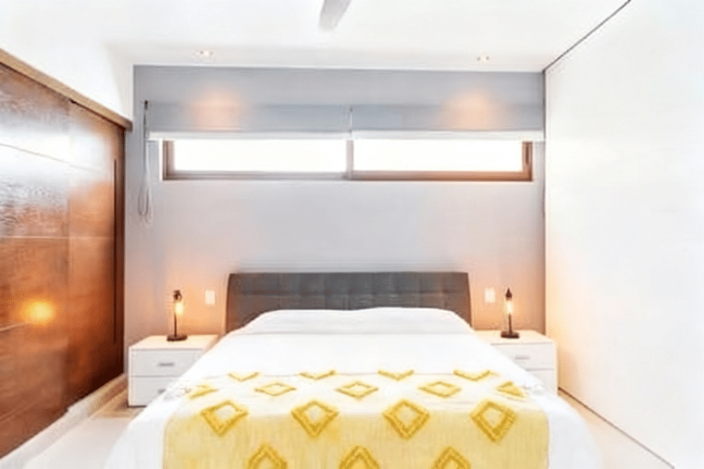 Bedroom with white walls, a bed with a yellow geometric-patterned cover, wooden nightstands, and a narrow horizontal window in a condo at Tao Tse M8 1-Bedroom Condo in Akumal.