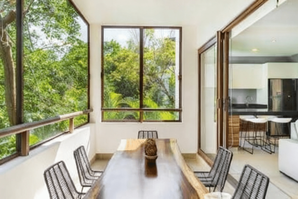 Bright dining area with large windows overlooking green trees and a view of the kitchen in the background at Tao Tse M8 at Tao Tse M8 1-Bedroom Condo in Akumal.