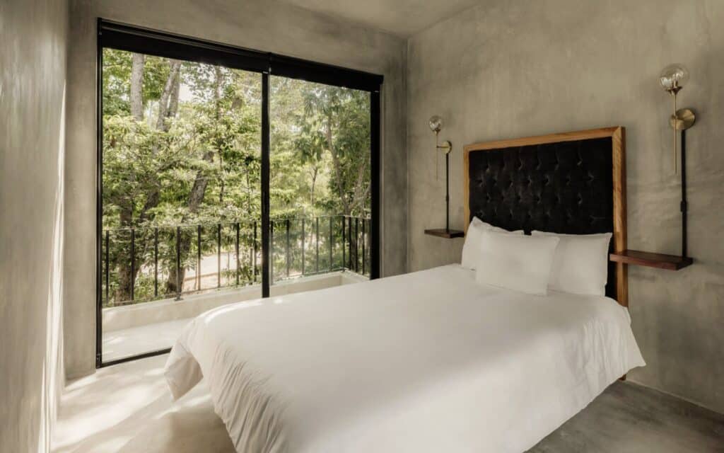 Bedroom with large window overlooking lush green trees and a balcony in Villa Asha at property name in area in Aldea Zama, Tulum.