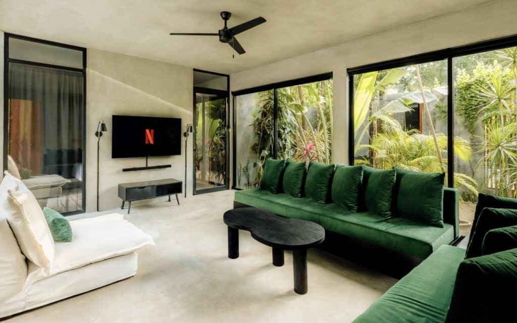 Living room with large windows showing lush greenery outside, modern white and green furniture, flat-screen TV, and a ceiling fan at Villa Asha in Aldea Zama, Tulum.