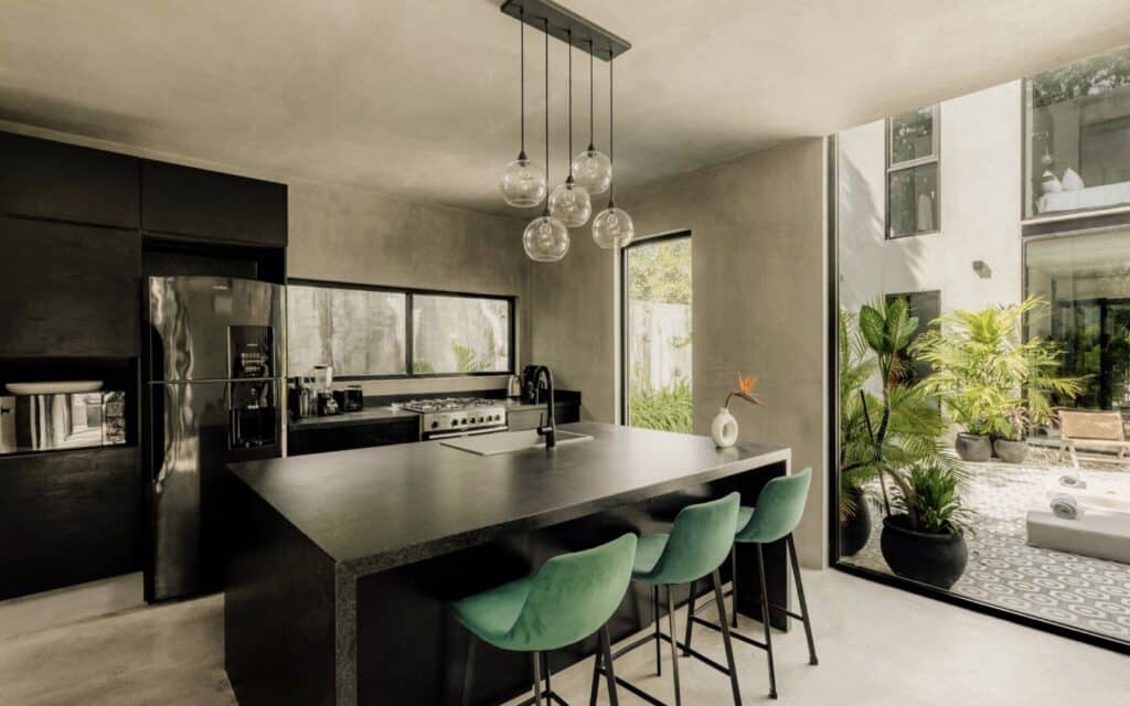 Modern kitchen with black cabinetry, large island, stainless steel appliances, and an adjacent dining area with green chairs, overlooking a garden in Aldea Zama, Tulum.
