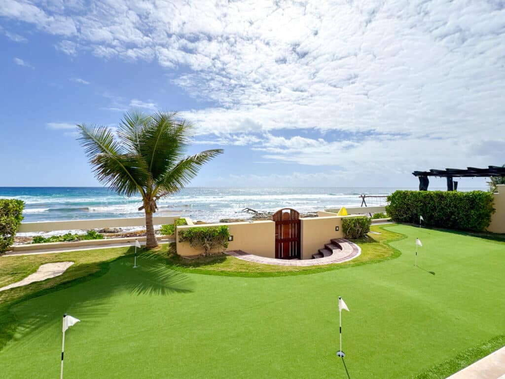 Villa Coccoloba in Tulum features a beachfront view with a palm tree, a wall with a gate, and a green putting area overlooking the ocean.