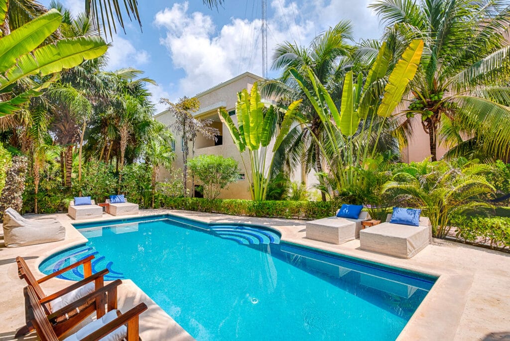 Villa Coccoloba in Tulum features a backyard with a rectangular swimming pool, surrounded by tropical palm trees and outdoor seating.