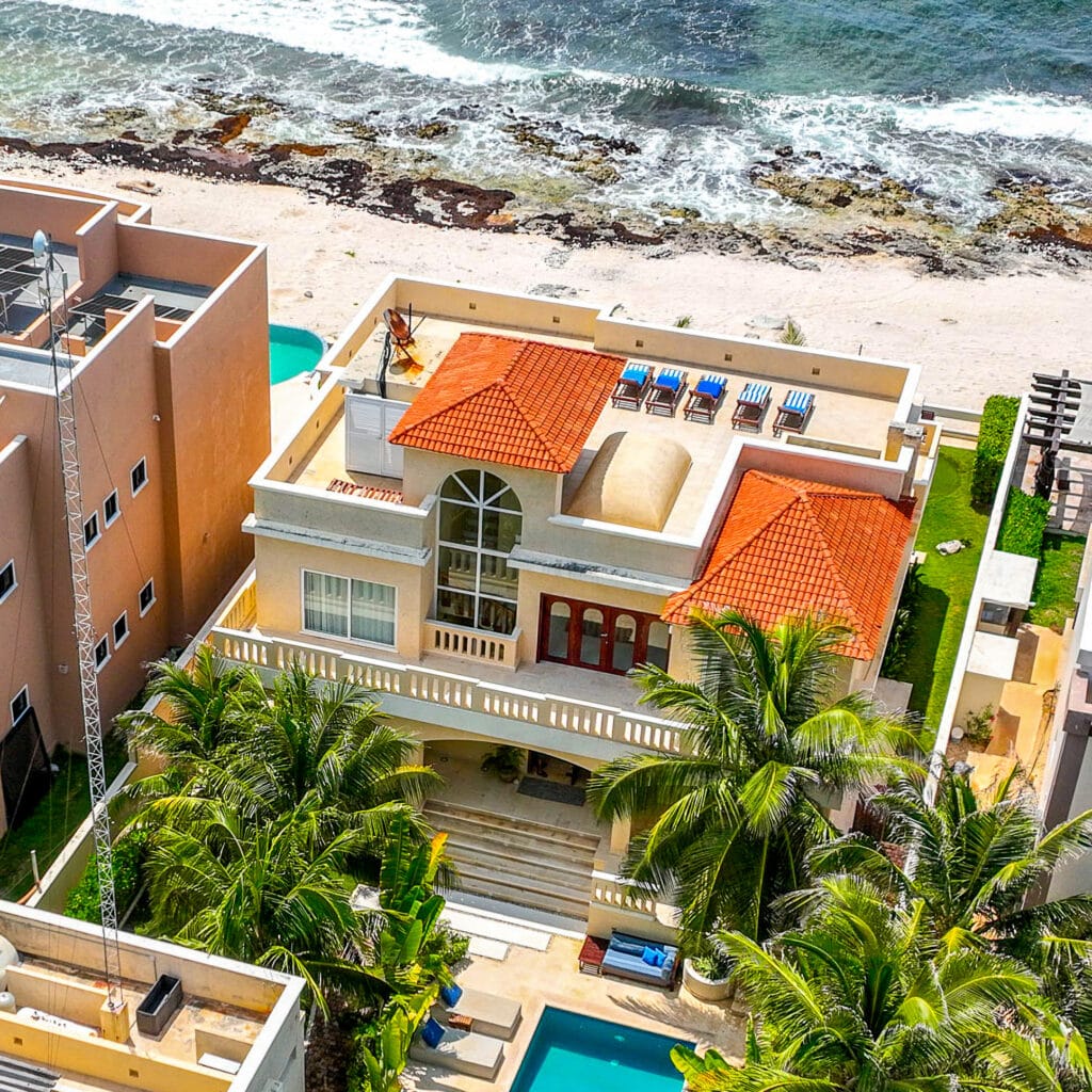 Aerial view of Villa Coccoloba in Tulum, featuring a beachfront property with a red-tiled roof, large arched window, a rooftop terrace with sun loungers, and lush palm trees.