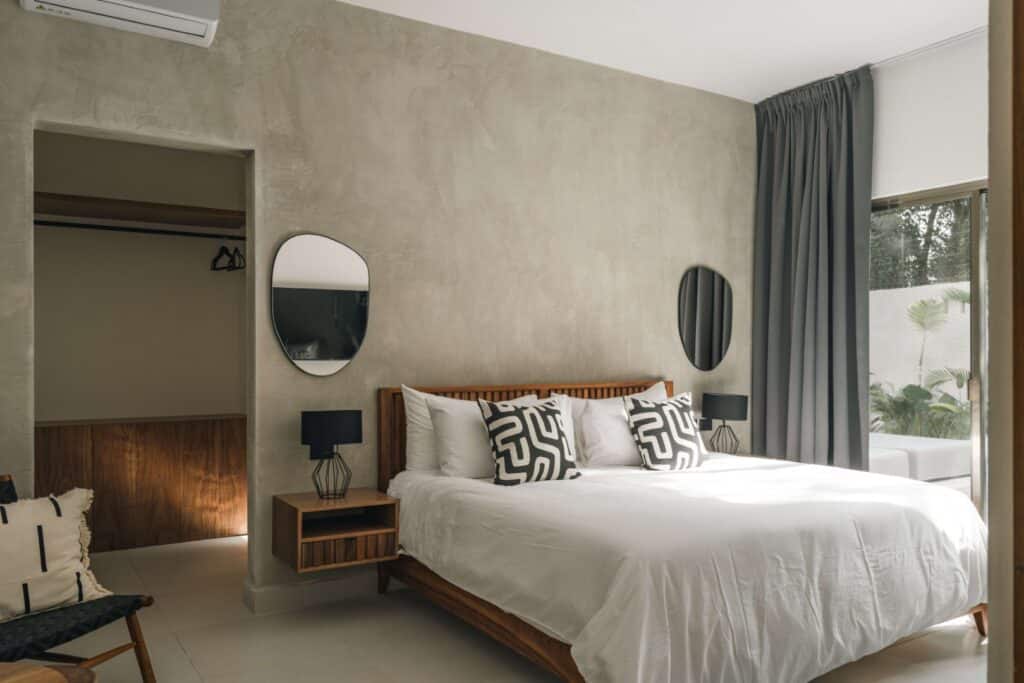 Bedroom with a wooden bed frame, white bedding, black and white pillows, and window with gray curtains at Villa Esperia in Aldea Zama, Tulum.