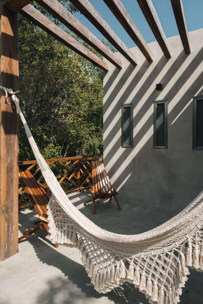 Wooden balcony with hammocks and chairs, cast shadows from pergola on gray wall, surrounded by trees at Villa Esperia in Aldea Zama, Tulum.