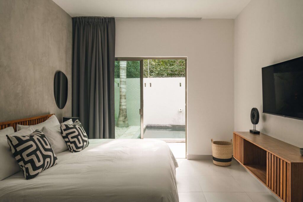 Bedroom with sliding glass door opening to an outdoor space, featuring white walls, wooden furniture, and modern decor at Villa Esperia in Aldea Zama, Tulum.