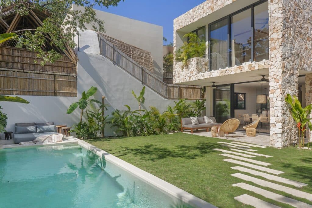 Modern house with large glass windows, stone exterior, outdoor seating, lush greenery, and a swimming pool in lush garden at Aldea Zama, Tulum at Villa Eterea.