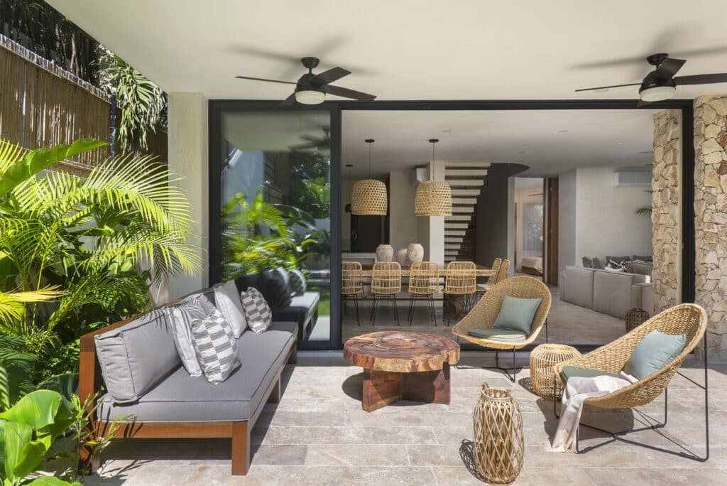 Covered outdoor living area with gray sofa, rattan chairs, lush tropical plants, and sliding glass doors leading to interior at Villa Eterea in Aldea Zama, Tulum.