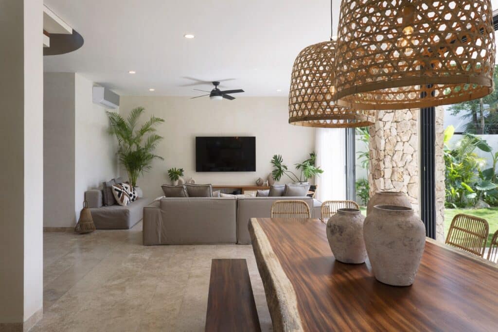 Living room with neutral-colored sofa, television on wall, indoor plants, and woven pendant lights near sliding glass doors to a garden at Villa Eterea in Aldea Zama, Tulum.