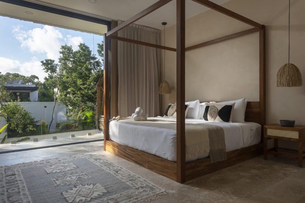 Bedroom with a wooden four-poster bed, sliding glass door opening to greenery, beige walls, hanging lamps, and a patterned rug at Villa Eterea in Aldea Zama, Tulum.