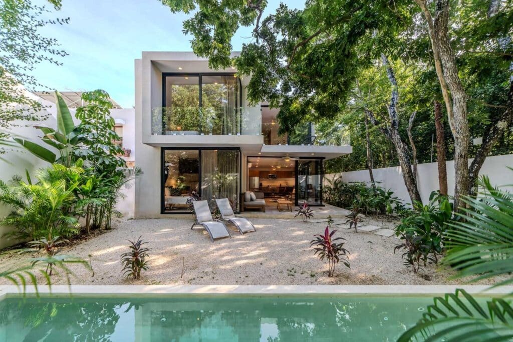 Modern two-story house with large glass windows and a backyard with lounge chairs, surrounded by lush trees at Villa Las Almas in Aldea Zama, Tulum.