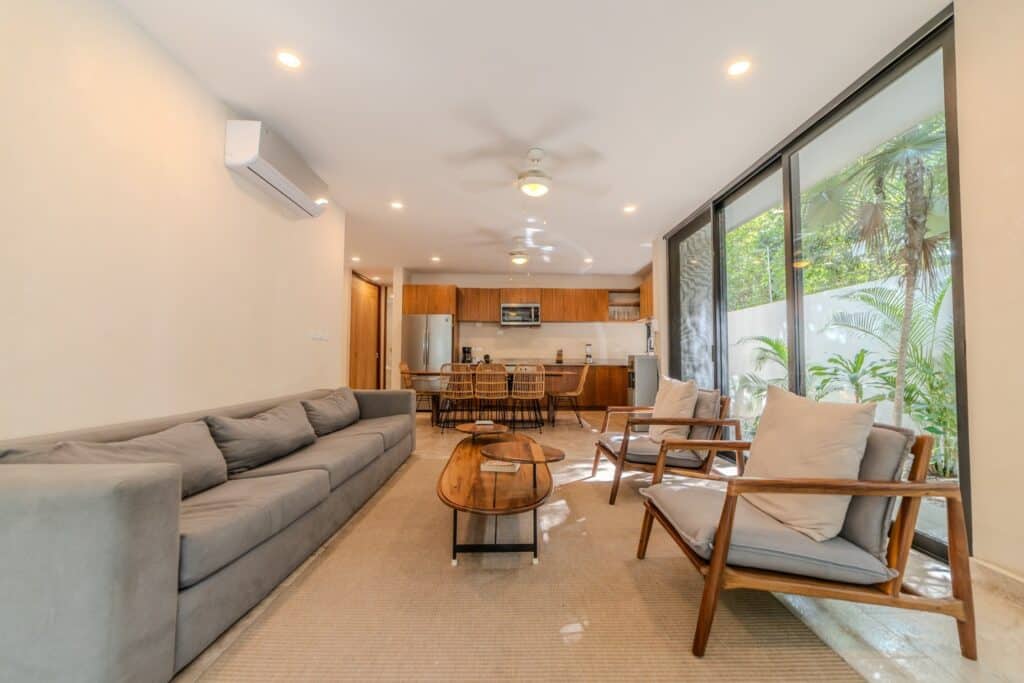 Living room with gray sofa, wooden chairs, glass sliding doors showing outdoor greenery, and open kitchen at Villa Las Almas in Aldea Zama, Tulum.